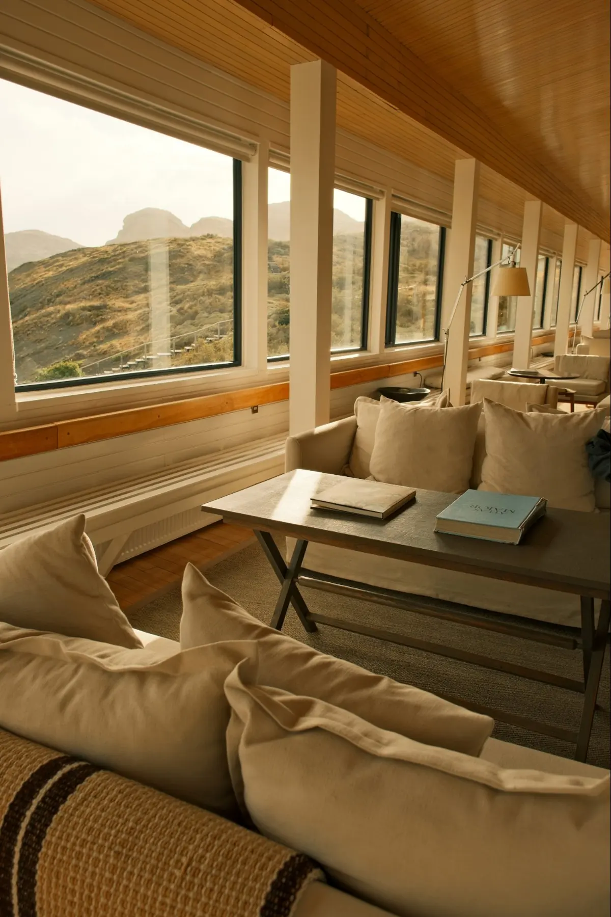 Linen sofas and books in a long, wood-ceilinged lounge lined with windows over the hills